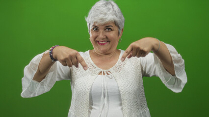 Woman, senior plus size with grey hair, points both index fingers downward while smiling, wearing white blouse and bracelets in studio; playful invitation.