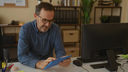 Senior hispanic man in glasses using tablet in modern office setting with organized desk, surrounded by computer, stationery, and shelves for business productivity and focus.