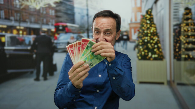 Man holding israeli shekel banknotes, standing in an urban city street with festive decorations, expressing happiness outdoors.