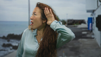 Woman cupping ear with hand and grimacing while listening on a seaside street pier by rocky shoreline and concrete promenade; confusion seeking clarity.