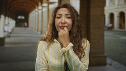 Fototapeta premium Woman touching chin with frown and hand near mouth on historic street arcade, striped yellow shirt visible; quiet concern.