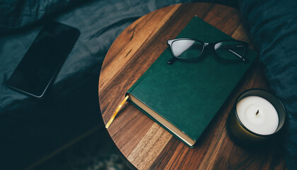 Green notebook and glasses rest on a wooden side table next to a candle and a smartphone at home during evening hours