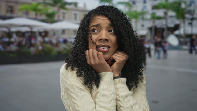 Woman pressing hands to cheeks with worried expression and gazing upward in a crowded city street; anxiety.