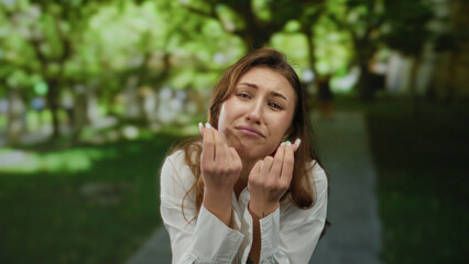 Young woman with playful expression gestures by pinching fingers together in a sunny outdoor park surrounded by greenery and trees, wearing a white shirt, conveying a humorous moment.
