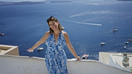 Naklejka premium Woman smiling with arms out on building terrace overlooking sea and boats in santorini town; vacation relaxation joy.