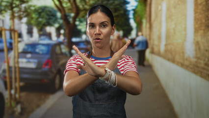 Woman in red striped shirt and denim apron with crossed arms and visible hands making an x gesture on a busy city street; refusal protest.