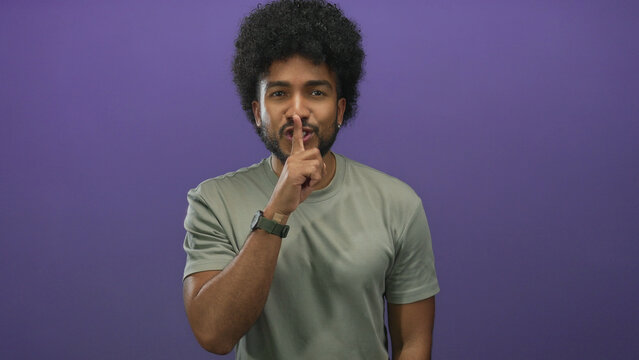 Man with curly hair in green shirt gesturing silence with finger on lips against a vibrant purple background, conveying secrecy and discretion in a studio setting.