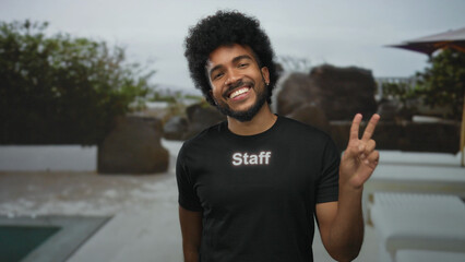 Man with afro smiling in staff uniform outdoors at hotel pool resort showing peace sign surrounded by greenery and white furniture capturing friendly hospitality vibe
