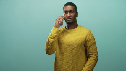 Young black man wearing glasses and yellow sweater, holding smartphone to ear and raising right...
