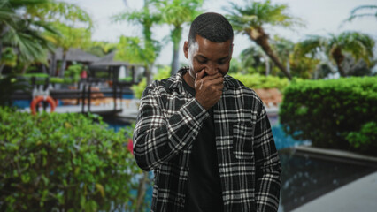 Young black man covering mouth with hand while laughing beside a pool and palm trees in front of a...