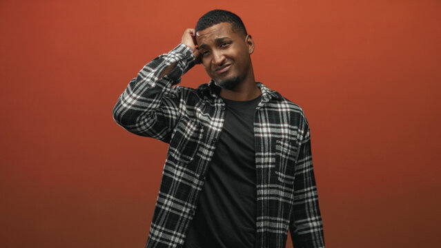 Young black man rubbing head with hand, wearing plaid shirt in studio against orange backdrop, eyes closed and wincing in discomfort; uncertainty reflection.