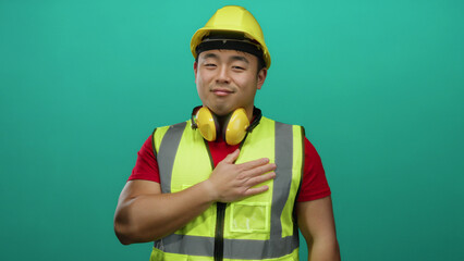 Man with yellow helmet and safety vest expressing gratitude against green wall background