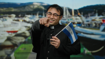 Young priest holding el salvador flag, smiling at seaside port with boats in background suggesting outdoor maritime location associated with travel and culture.