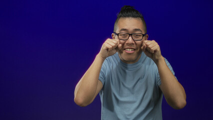 Obraz premium Man with clenched fists near face showing hands and forearms, wearing glasses and light blue t shirt in deep blue studio; joy simple expression.