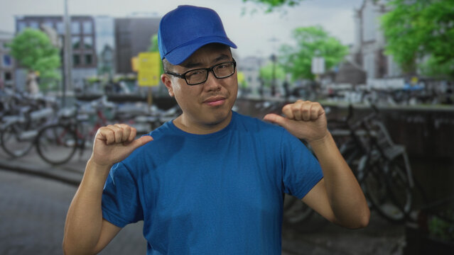 Man in blue cap and t shirt points both thumbs to his chest on a busy street in amsterdam with bicycles and canal visible; self promotion confidence. - Powered by Adobe