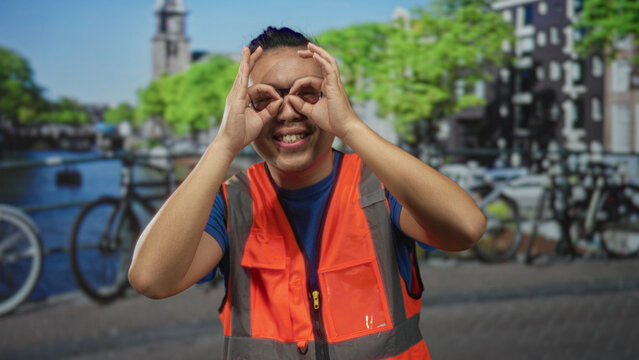 Young man in orange safety vest making hand binoculars on a street in amsterdam; playful curiosity exploration.