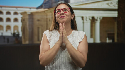 Woman smiling with hands pressed together wearing red glasses and white sleeveless top in front of building facade  gratitude. © Krakenimages.com