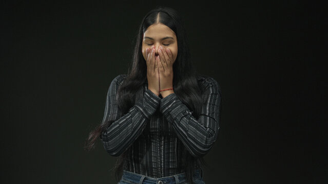 Young latin woman with hands covering mouth gesture in studio with dark green background; surprise.