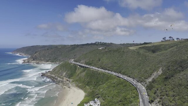 Drone flies west along coastal highway above beach as paragliders fill the sky on a sunny day on the Garden Route in Wilderness, South Africa