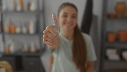 Teenage girl artisan holds a clay coated thumb for a thumbs up in a pottery studio lined with shelves; optimism.