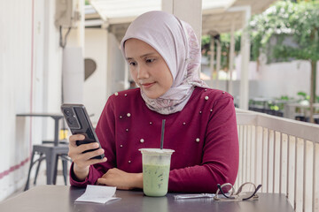 An Asian Muslim woman replying to messages on her smartphone while sitting in a coffee shop