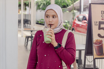 A young Indonesian Muslim woman standing while enjoying her iced matcha drink