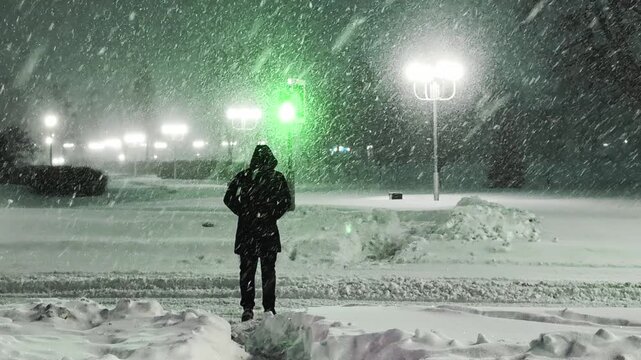 Person walks across snow-covered pedestrian crossing on city street at night. Heavy snow falls, illuminated by streetlights and changing traffic signal. Hooded figure navigates urban path