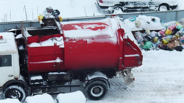 Waste collection vehicle processing garbage bags in winter environment, heavy snow blankets street and surroundings, difficult conditions for sanitation service, accumulation of trash bags near site