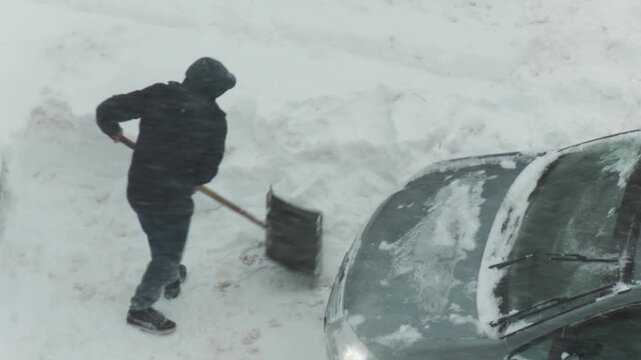 Overhead view person shoveling fresh heavy snow from around passenger car in winter. Vehicle stuck in deep snowdrift requiring effort to clear path. Cold weather conditions and hard work apparent.