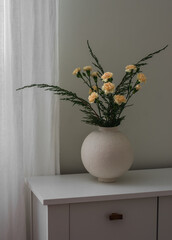 A bouquet of juniper branches and yellow carnations in a round ceramic vase on a white chest of drawers in the living room
