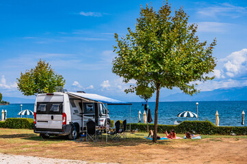 A scenic summer day at a Lake Ohrid campsite in Udenisht, Albania, featuring a camper van with an awning and a family sunbathing on the grass under a tree with clear blue waters in the background.