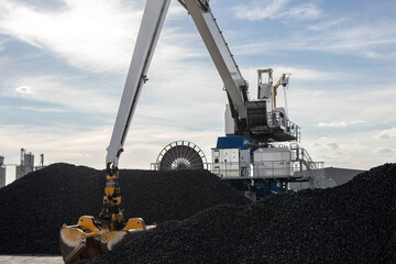 Large industrial crane loads dark coal piles bright sunny sky  backlight background. Metallic blocks and massive equipment storage site. Energy commodity industry global trade logistics business