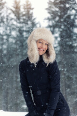 Portrait young adult mid age smile woman enjoy have fun walking in park forest snowfall cold winter day. Female portrait person happiness wear black ski overall stylish fur hat snowflakes background