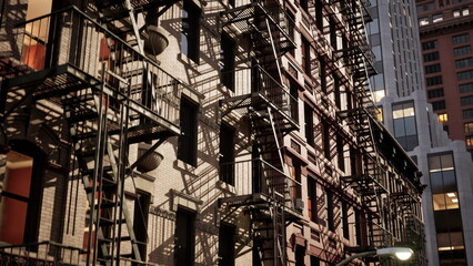 Naklejka premium Late evening light casts intricate shadows from fire escapes onto a historic building. The vibrant city skyline looms above, showcasing modern architecture alongside classic designs.