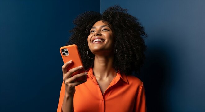 Joyful young black woman with an afro hairstyle smiling while using her smartphone against a dark blue background.
