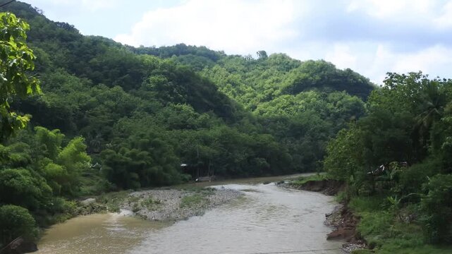 View of the Oyo River in Kedungjati, Selopamioro, Imogiri, Yogyakarta, Indonesia, flanked by two hills with lush green plants growing around it with a bright blue sky in the background during the day