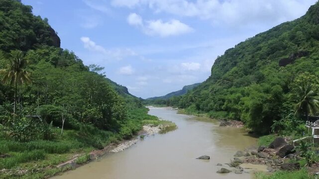 View of the Oyo River in Kedungjati, Selopamioro, Imogiri, Yogyakarta, Indonesia, flanked by two hills with lush green plants growing around it with a bright blue sky in the background during the day