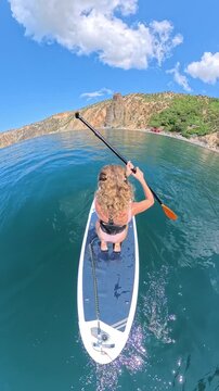 Woman paddleboard ocean, kneeling and paddling in clear blue water with scenic coastal landscape on a sunny day. Vertical video