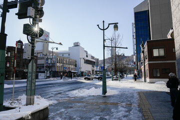 A wide-angle, realistic photograph captures a sun-drenched winter street scene at an intersection in a Japanese city, The asphalt and sidewalks are partially covered in snow and slush.