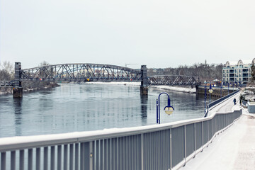 Snowy morning at Magdeburg Schleinufer shows calm river Elbe reflecting steel Hubbrucke bridge. Quiet winter path lined with blue lamps and benches creates serene urban landscape
