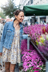 Young adult joyful woman choose to buy colorful potted flowers at outdoor market stall. Female person at sale of blooming plants in pots at botanical street German store on sunny spring autumn day