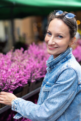 Young adult joyful woman choose to buy colorful potted flowers at outdoor market stall. Female person at sale of blooming plants in pots at botanical street German store on sunny spring autumn day