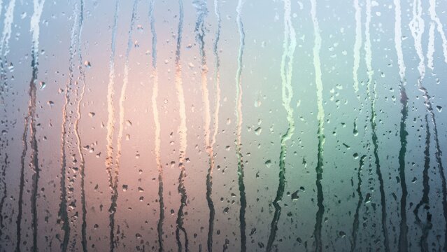 Abstract background of water droplets and streaks on wet window glass, with a blurred colorful light gradient creating a serene atmospheric texture.