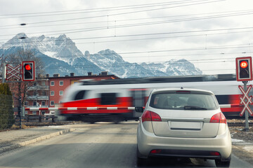 Car stopped railway crossing Austria fast train passes motion blur red signal lights active safety barriers down. Alpine mountains urban suburbs power lines overcast winter atmosphere