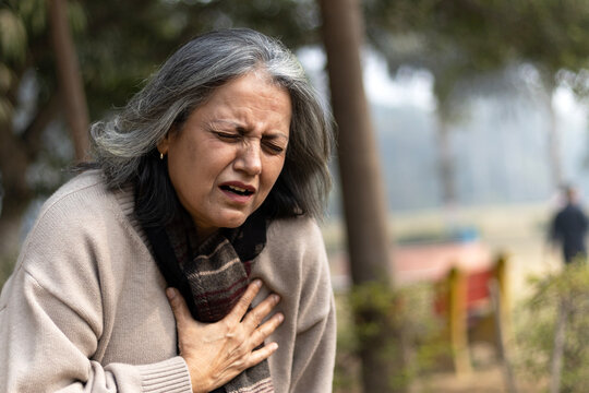 Indian senior woman holding chest with pained expression sitting on bench outdoors. Possible heart diseases problem, outdoor got troubles with her chest pain or breathing difficulty in park