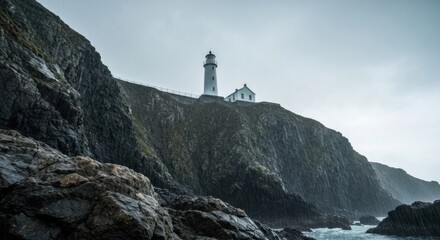 A stark white lighthouse stands atop a rugged, rocky cliff under a moody, overcast sky