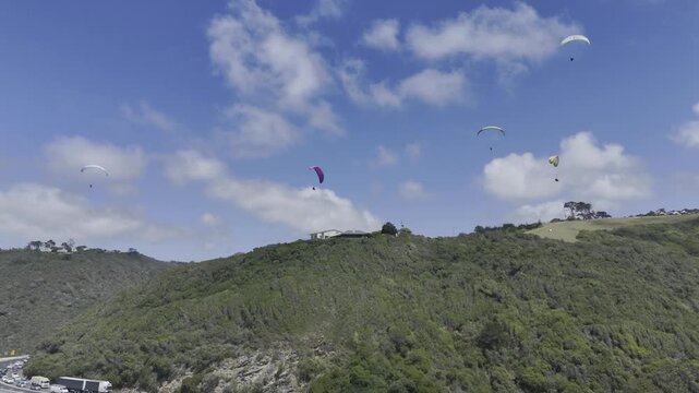 Drone flies to the right below a sky full of paragliders on a sunny day on the Garden Route in Wilderness, South Africa