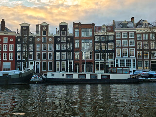 Daytime Amsterdam canal scene: historic Dutch houses and houseboats reflect on rippled water under a cloudy winter sky, authentic travel view