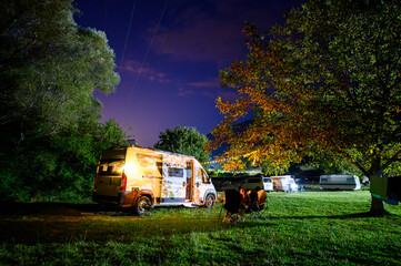 Night camping in nature near river Zeta, Montenegro, with a camper van, chairs, and warm lights under trees, capturing a cozy riverside camping atmosphere and van life experience.