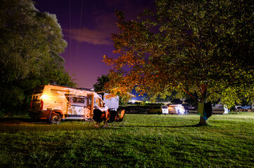 Night camping in nature near river Zeta, Montenegro, with a camper van, chairs, and warm lights under trees, capturing a cozy riverside camping atmosphere and van life experience.